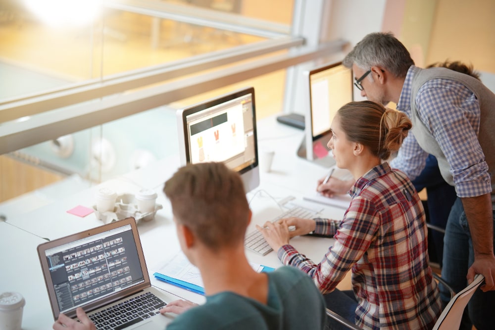 Drei Personen arbeiten an Computern in einem modernen Büro mit großen Fenstern.