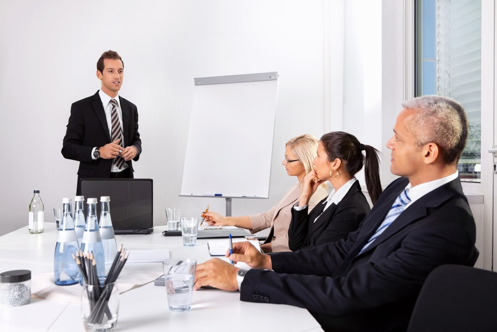 Ein Mann im Anzug hält eine Präsentation vor seinen 3 Personen in einem modernen Konferenzraum mit Flipchart.