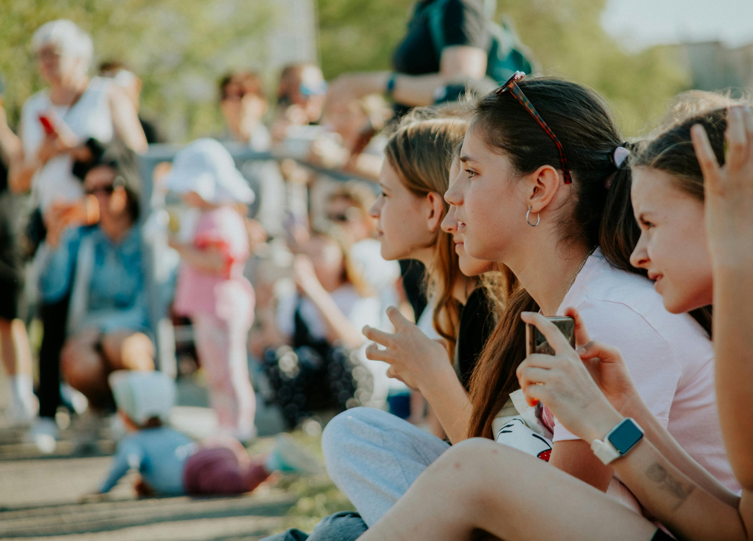 Eine Gruppe von Menschen ist zu sehen. Es scheint ein sonniger Tag zu sein. Im Vordergrund sitzen vier Mädchen.
