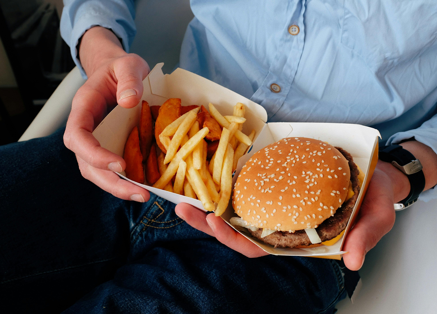 Eine Person in einem blauen Hemd und einer Jeanshose hält einen Bürger und Pommes in den Händen.
